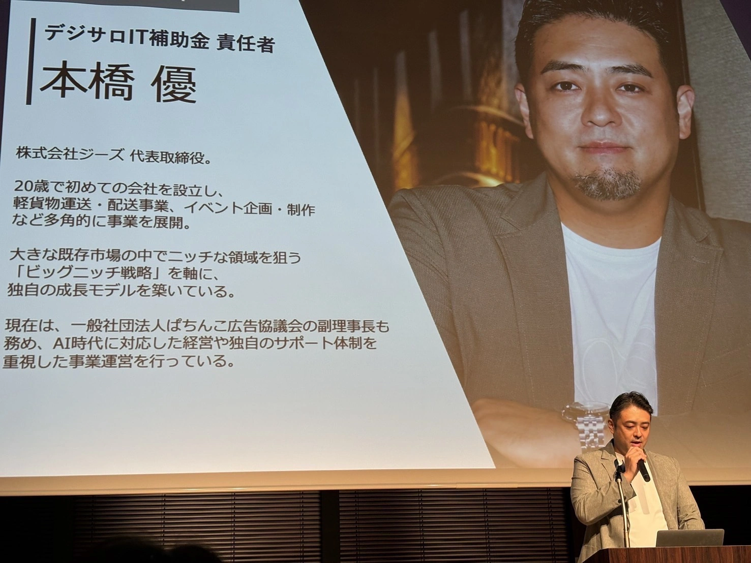 デジサロIT補助金責任者の本橋優氏が、プロフィール紹介スライドを背景に登壇している様子